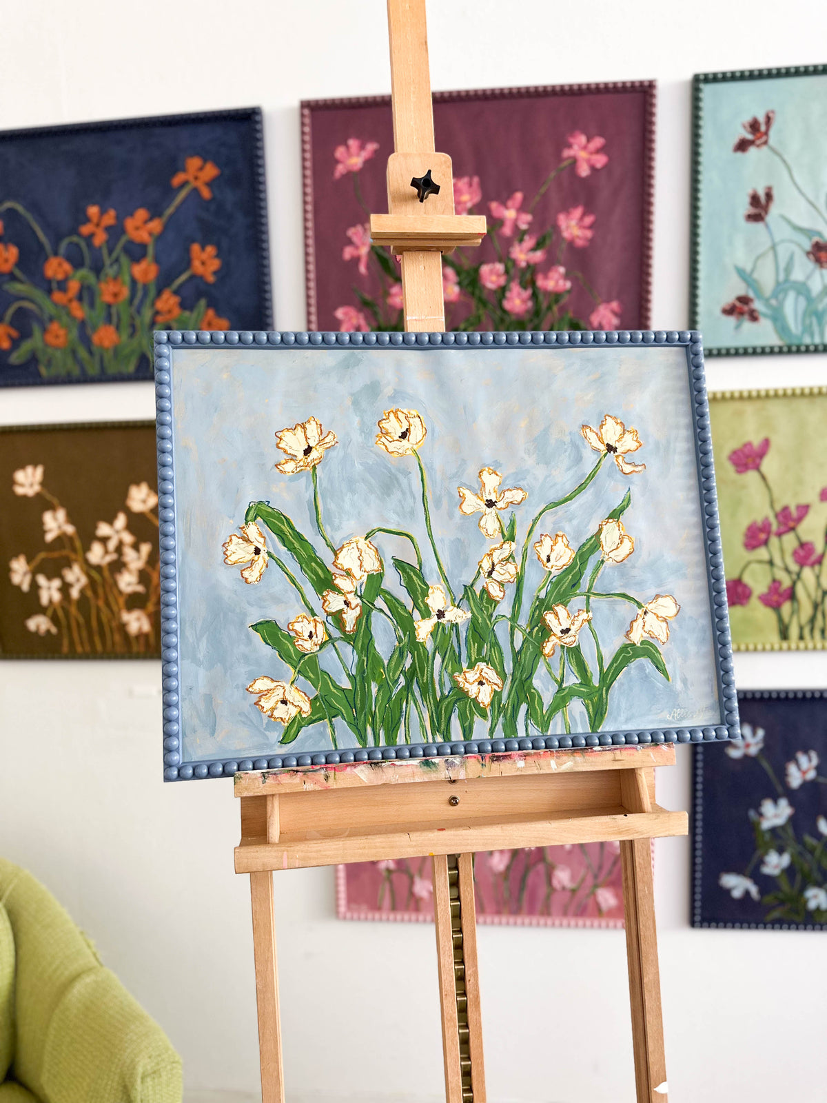 A painting of off white flowers with green leaves on a light blue background and framed in a blue bobbin frame sitting on an easel in from of other paintings.