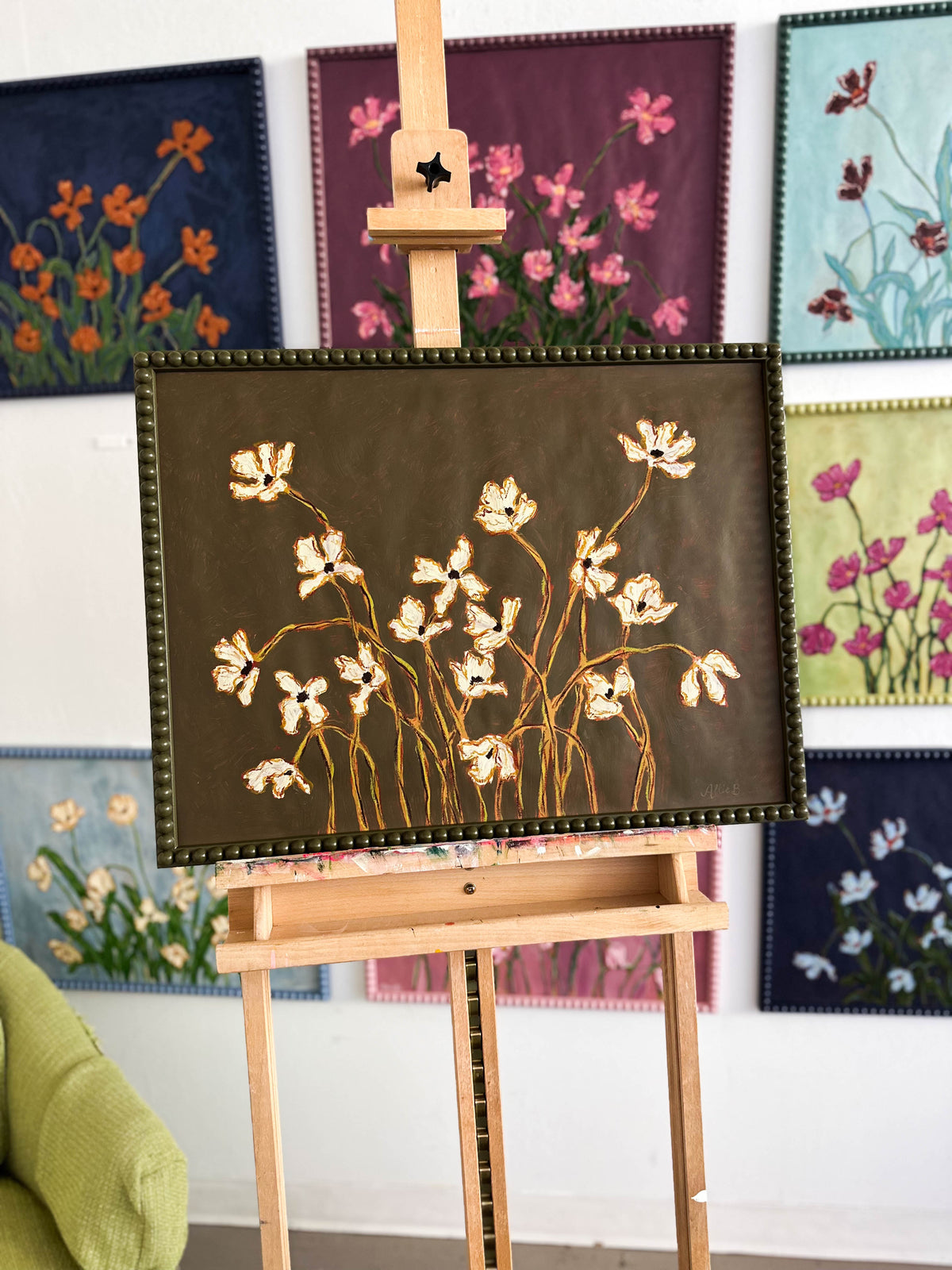 A painting of off white flowers with green stems on a thyme green background framed in a thyme green bobbin frame. The painting is on an easel in front of a wall full of paintings.