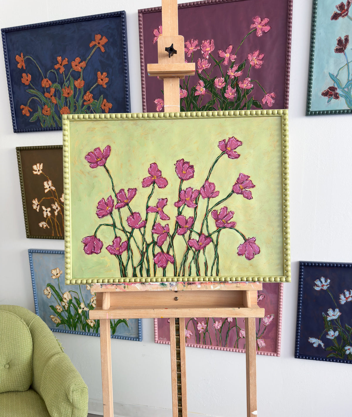 A painting of deep pink flowers with green stems on a chartreuse background framed in a chartreuse green bobbin frame. The painting is on an easel in front of a wall full of paintings.