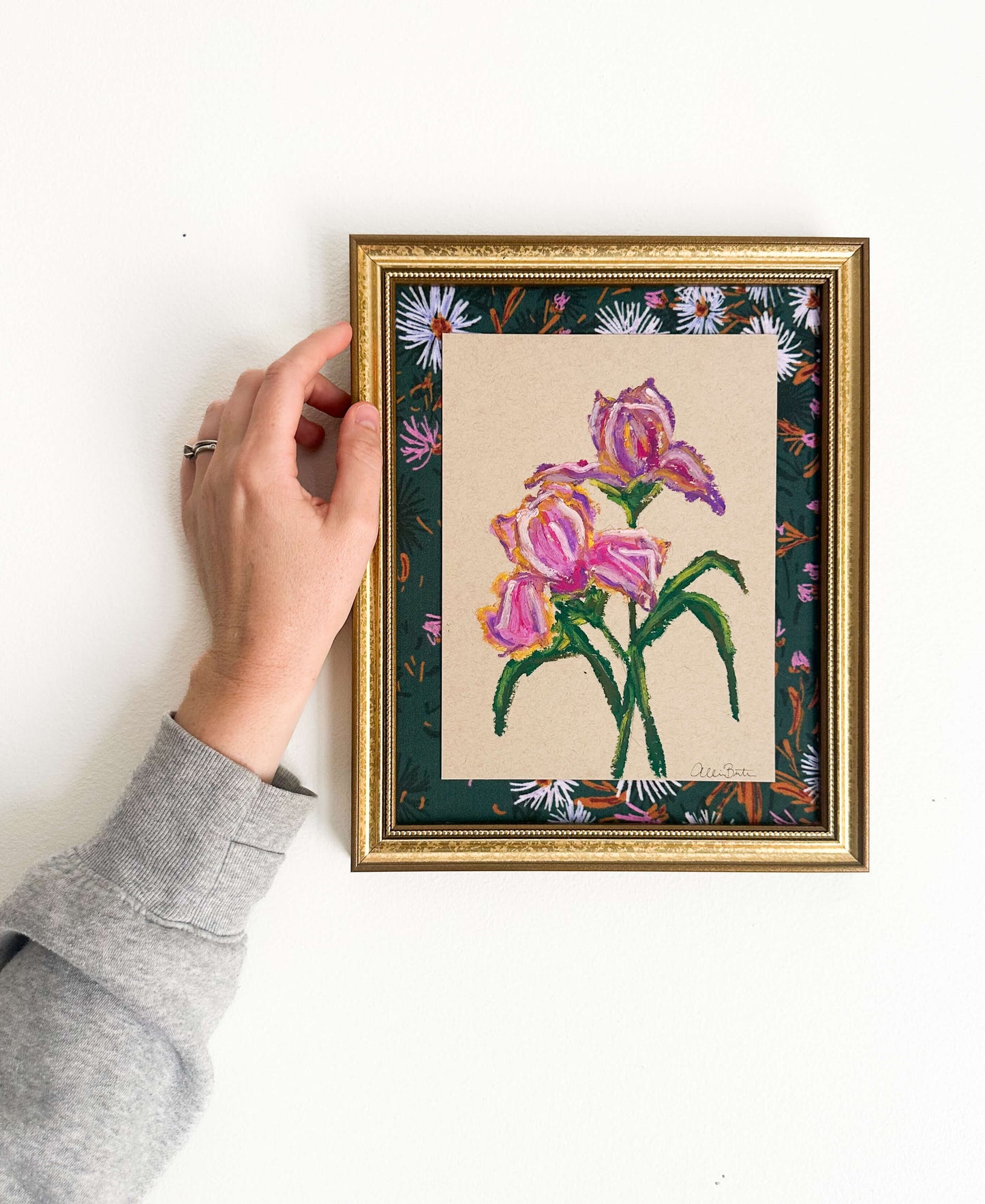 A painting of purple irises on tan paper mounted on dark floral fabric. The painting is in a gold frame and shown on a white wall.