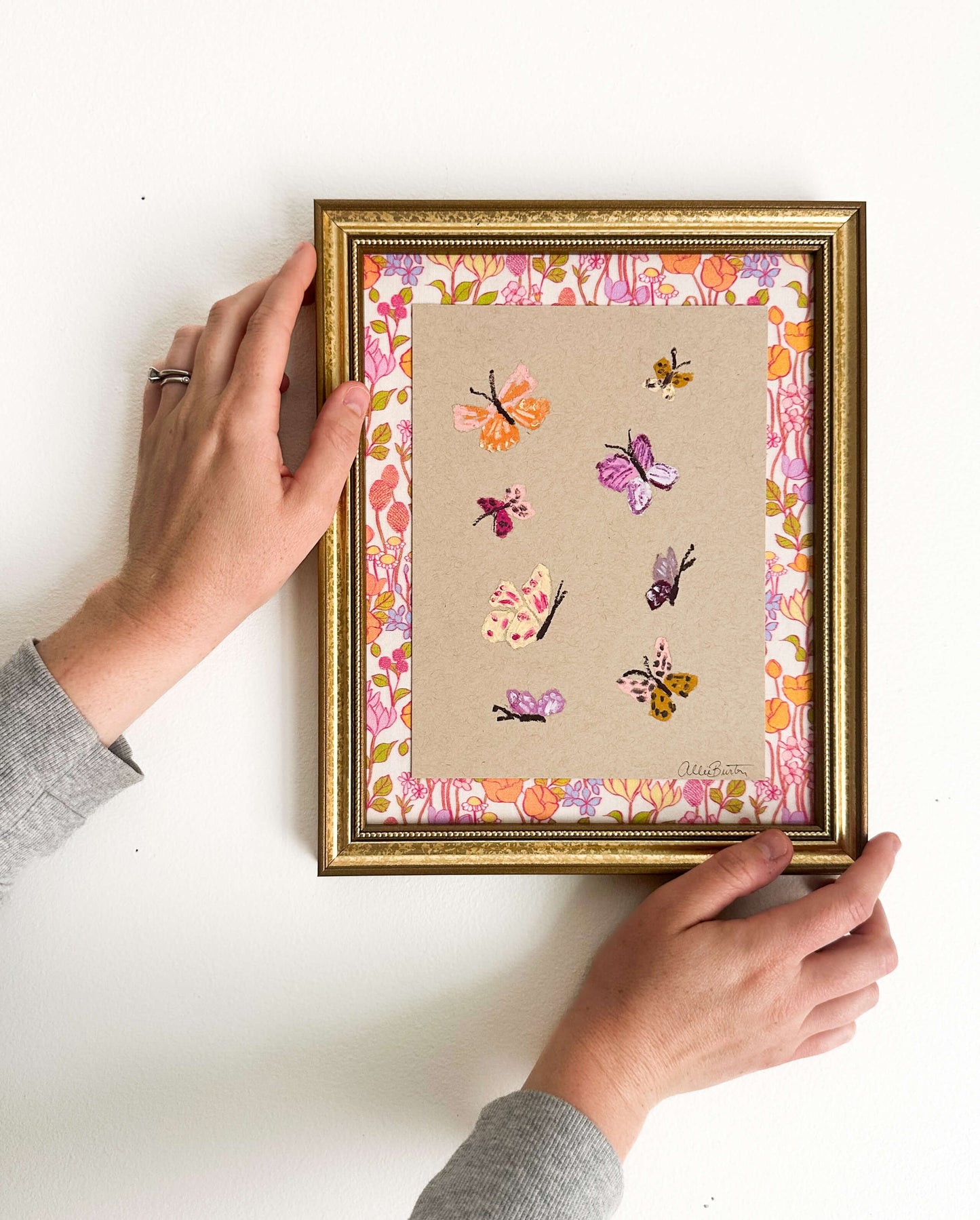 Eight colorful butterflies painted on tan toned paper and mounted on colorful, floral fabric. The painting is framed in a gold frame and shown on a white wall.