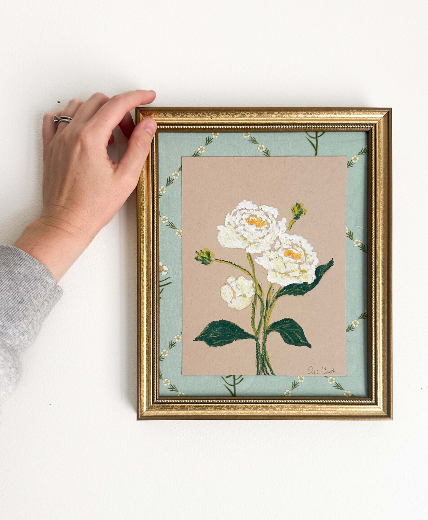 A painting of white peonies on tan paper mounted on floral fabric. The painting is in a gold frame and shown on a white wall.