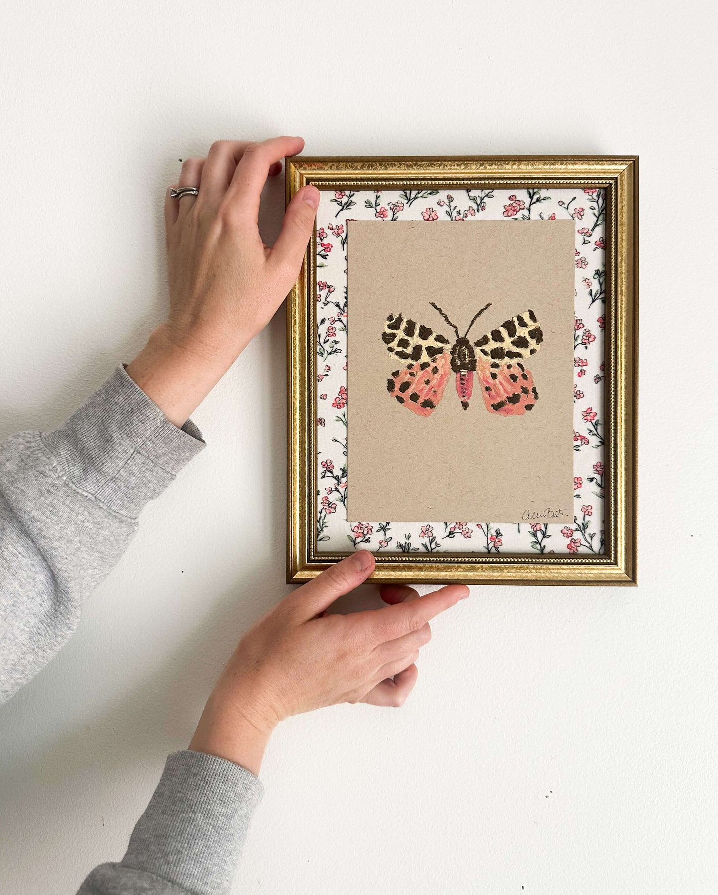 a pink, yellow and brown moth painted on tan paper. The paper is mounted on floral fabric, framed in a gold frame, and is shown on a white wall.
