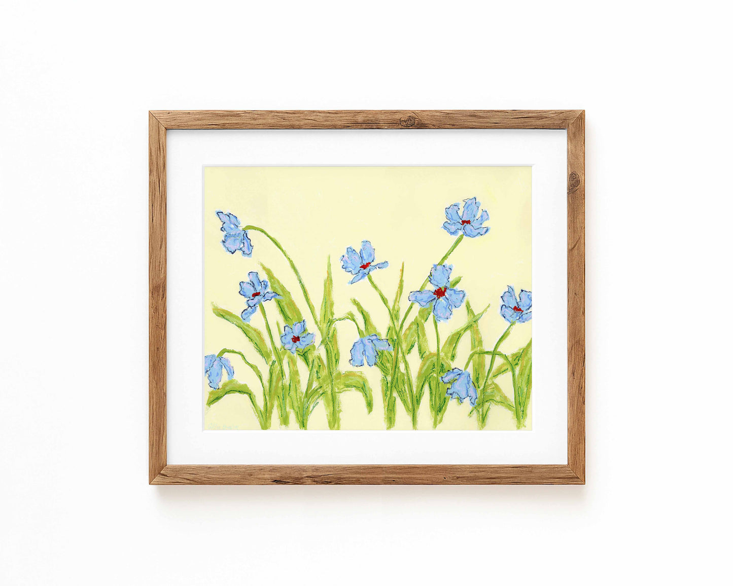 A framed print of blue irises on a pale yellow background, set in a light brown wooden frame with a white mat, displayed against a clean white wall.