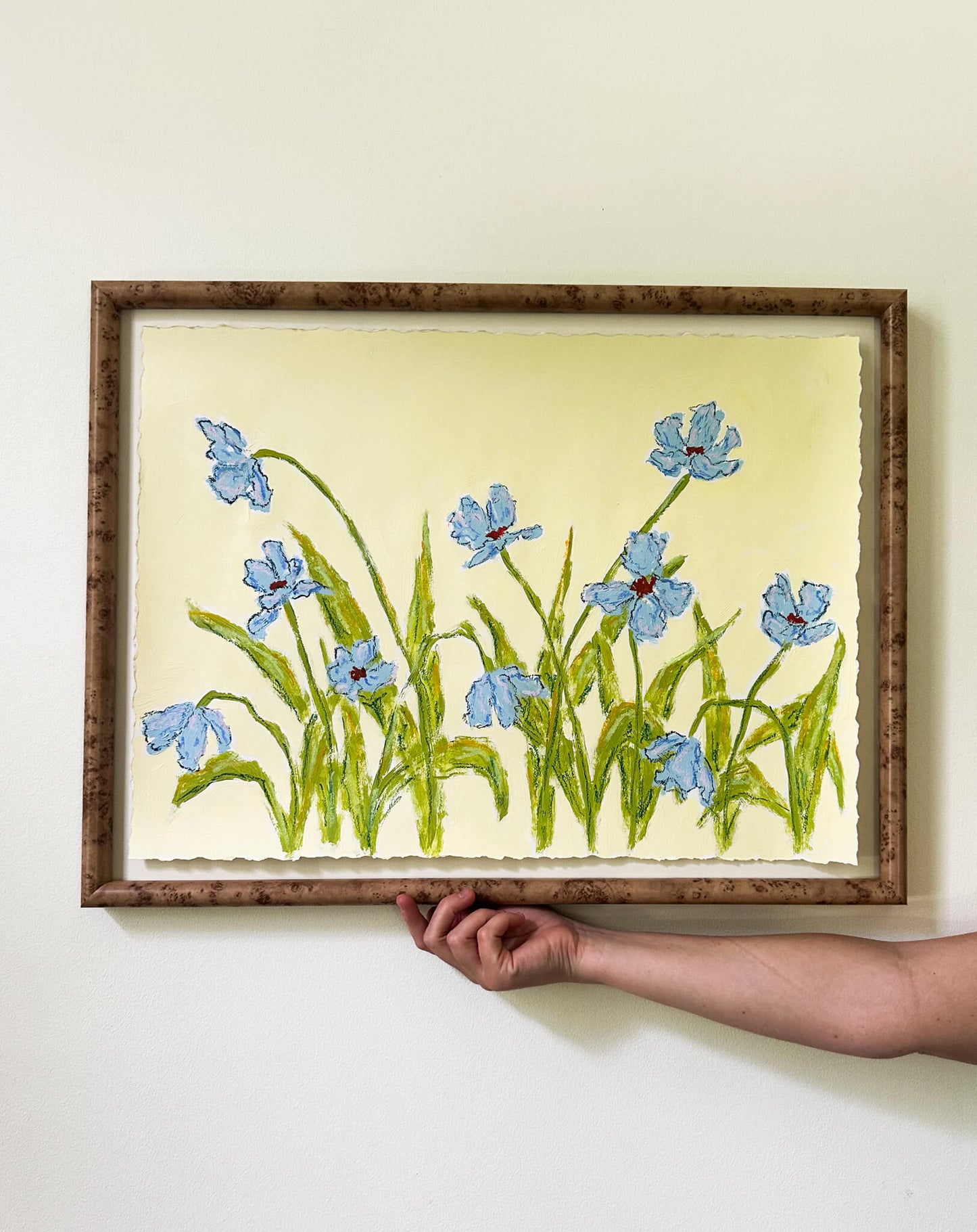 Framed embroidered artwork of blue flowers held by a hand against a white wall