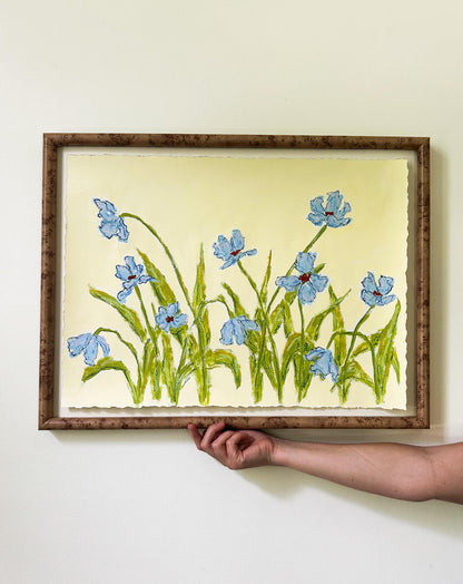 Framed embroidered artwork of blue flowers held by a hand against a white wall