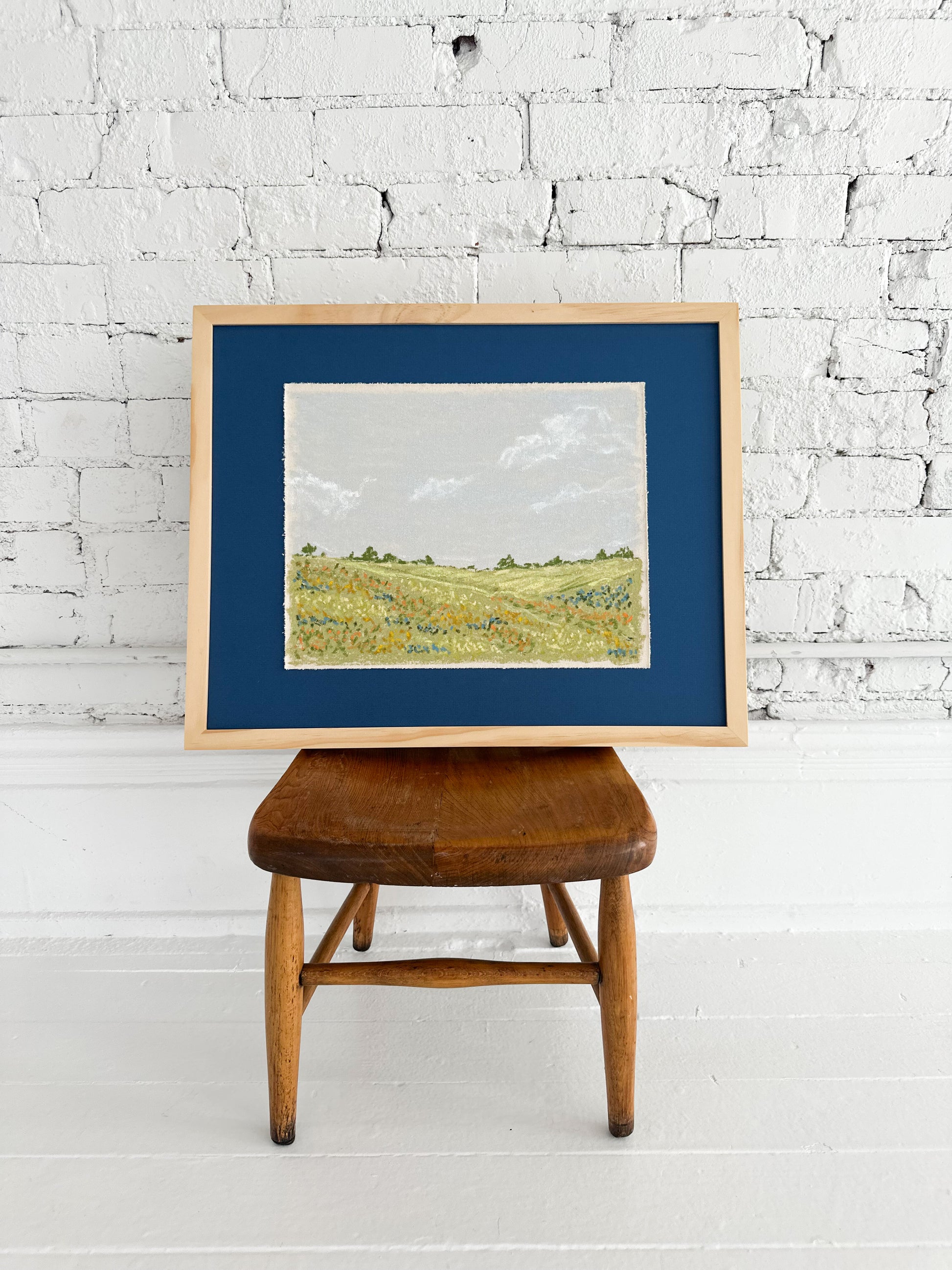 Framed landscape painting on a wooden stool against a white brick wall.