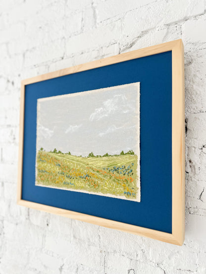 Framed artwork of a landscape with blue mat and frame on a white textured wall