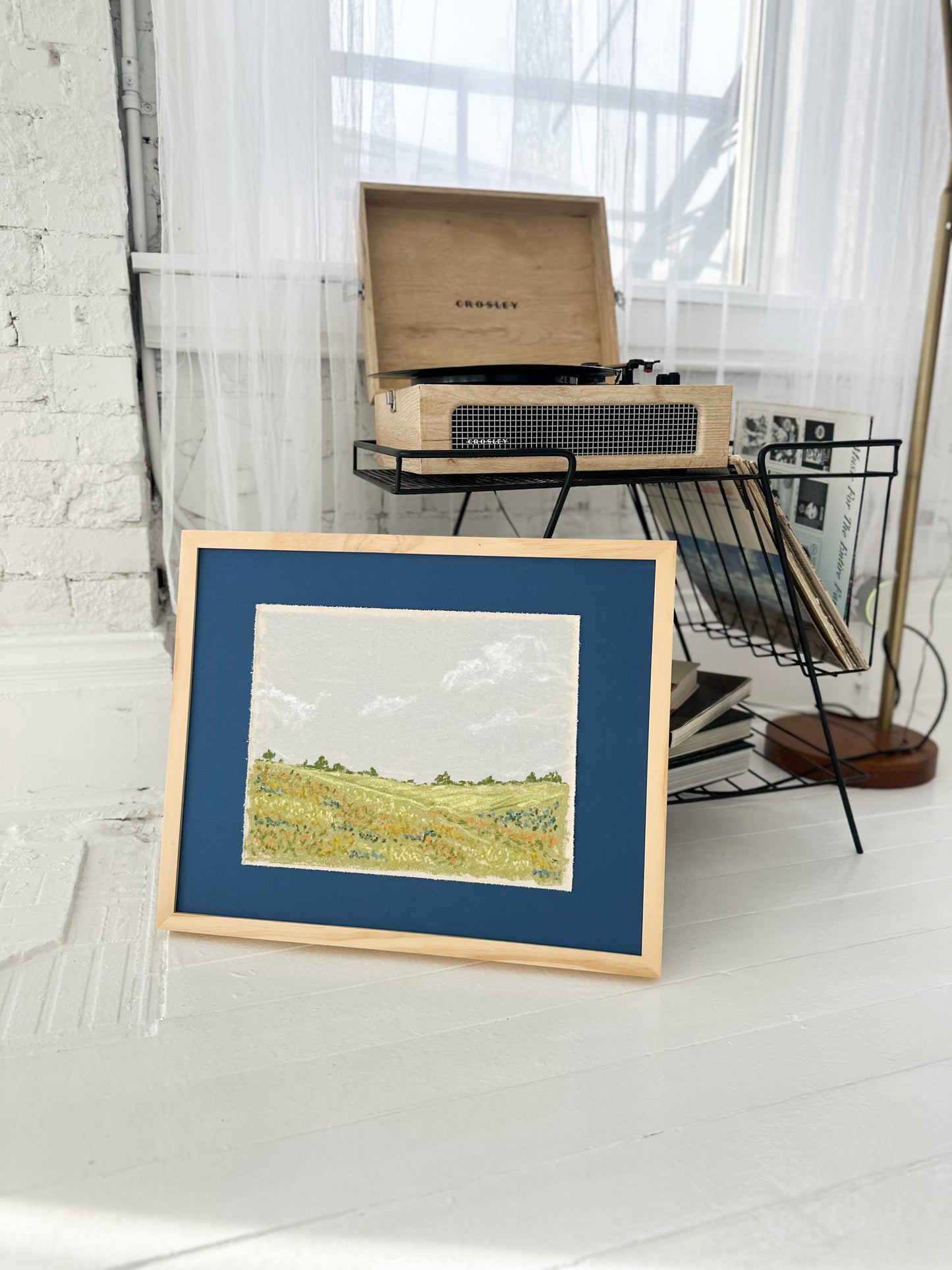Framed artwork of a landscape on a white floor with a record player in the background.