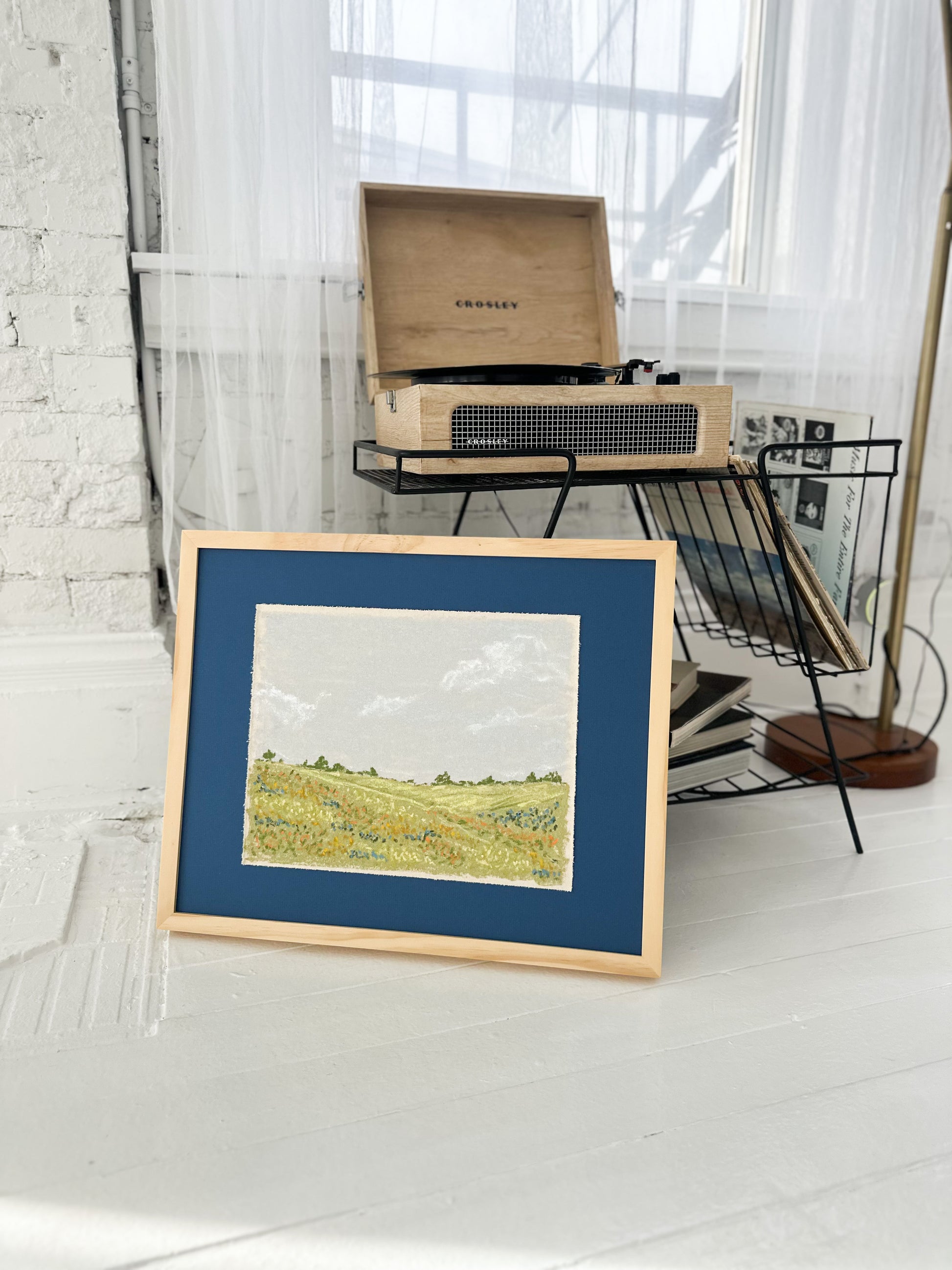 Framed artwork of a landscape on a white floor with a record player in the background.