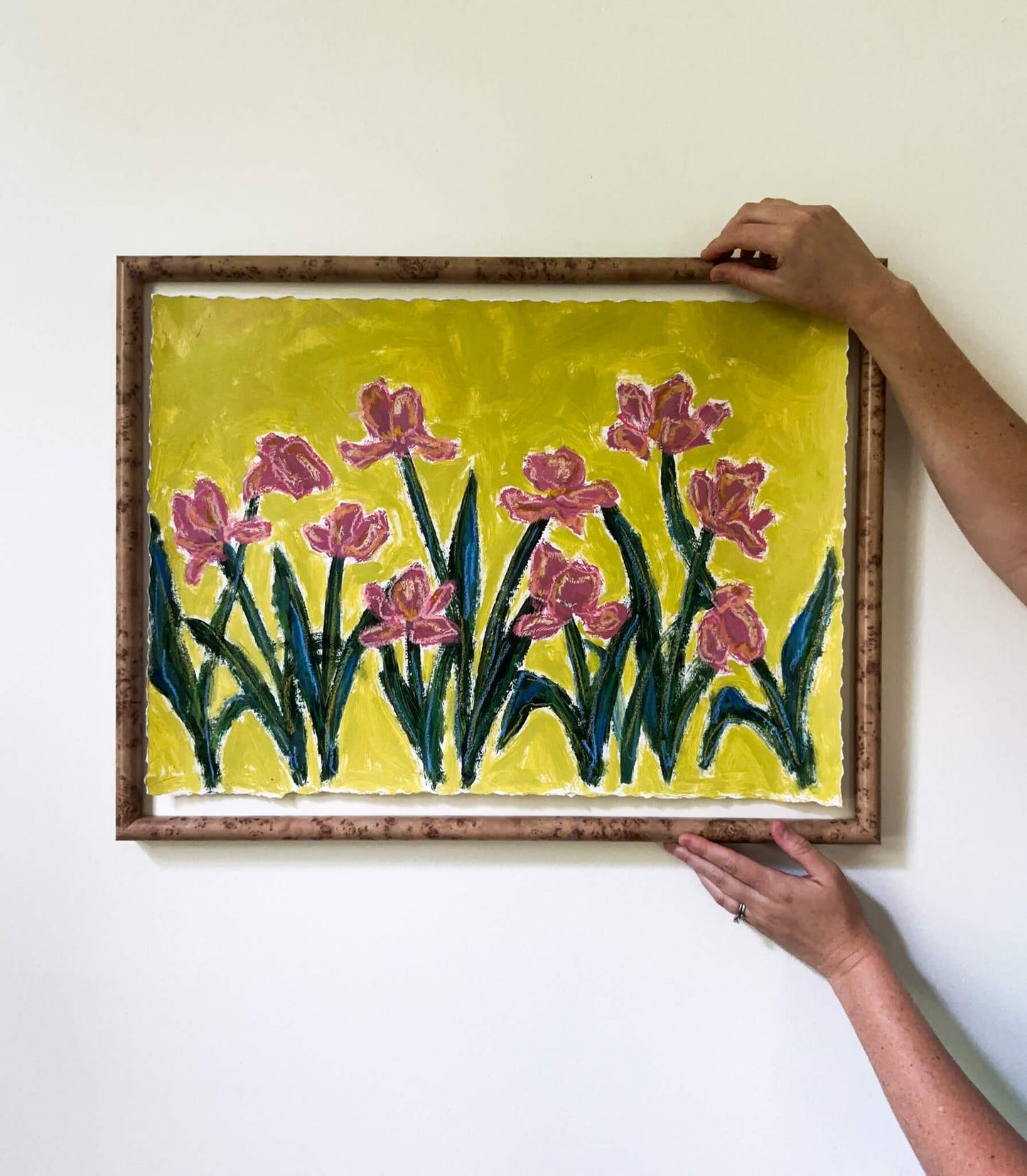 Framed artwork of pink flowers on a yellow background held by two hands.