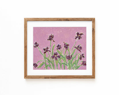 Framed floral wall art featuring purple irises on a textured pink background, set in a light natural wood frame with a white mat against a white wall.