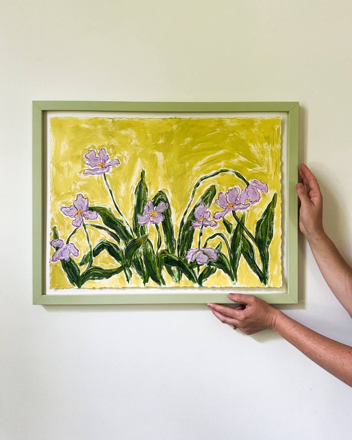 Framed painting of purple flowers on a yellow background, held by a person.