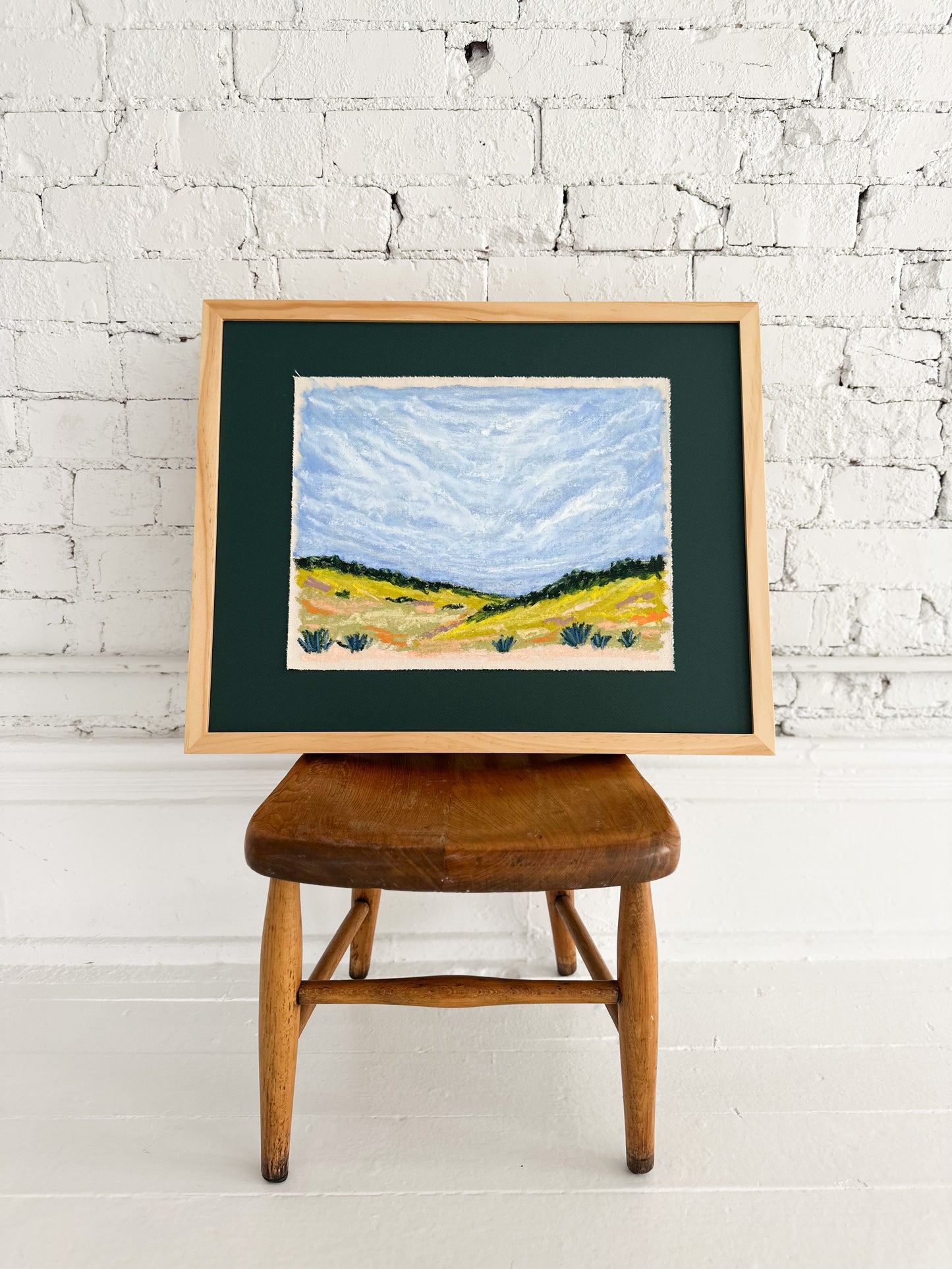 Framed landscape painting on a wooden stool against a white brick wall.