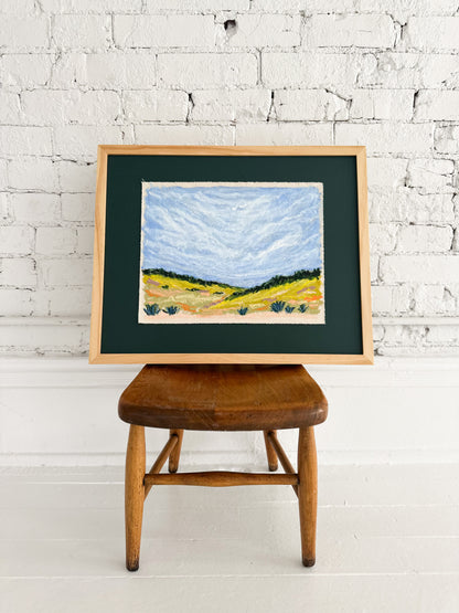 Framed landscape painting on a wooden stool against a white brick wall.