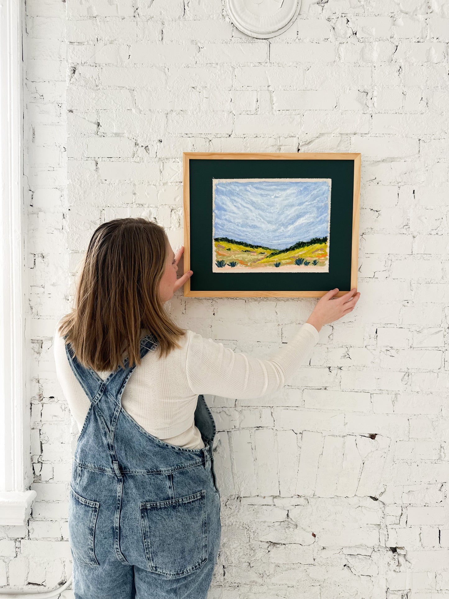 Person hanging a framed landscape painting on a white brick wall