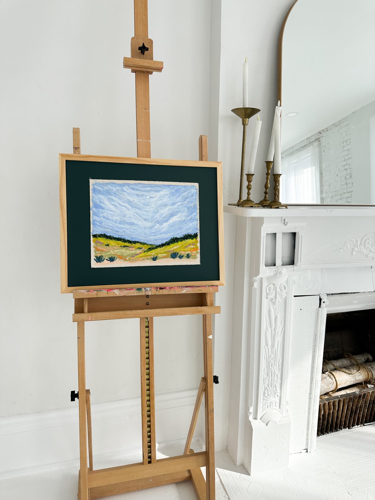 Painting on an easel in a room with a fireplace and mirror.