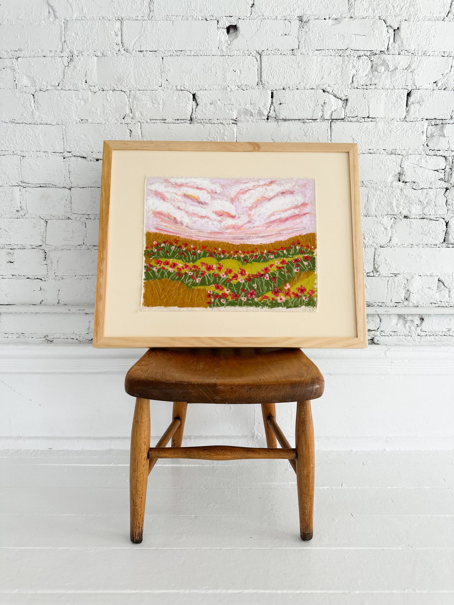 Framed artwork of a landscape with flowers on a wooden stool against a white brick wall.