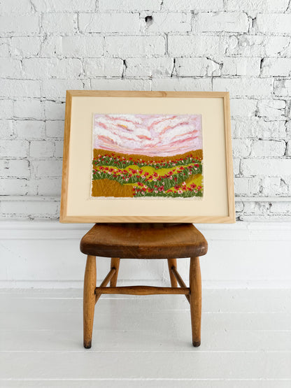 Framed artwork of a landscape with flowers on a wooden stool against a white brick wall.