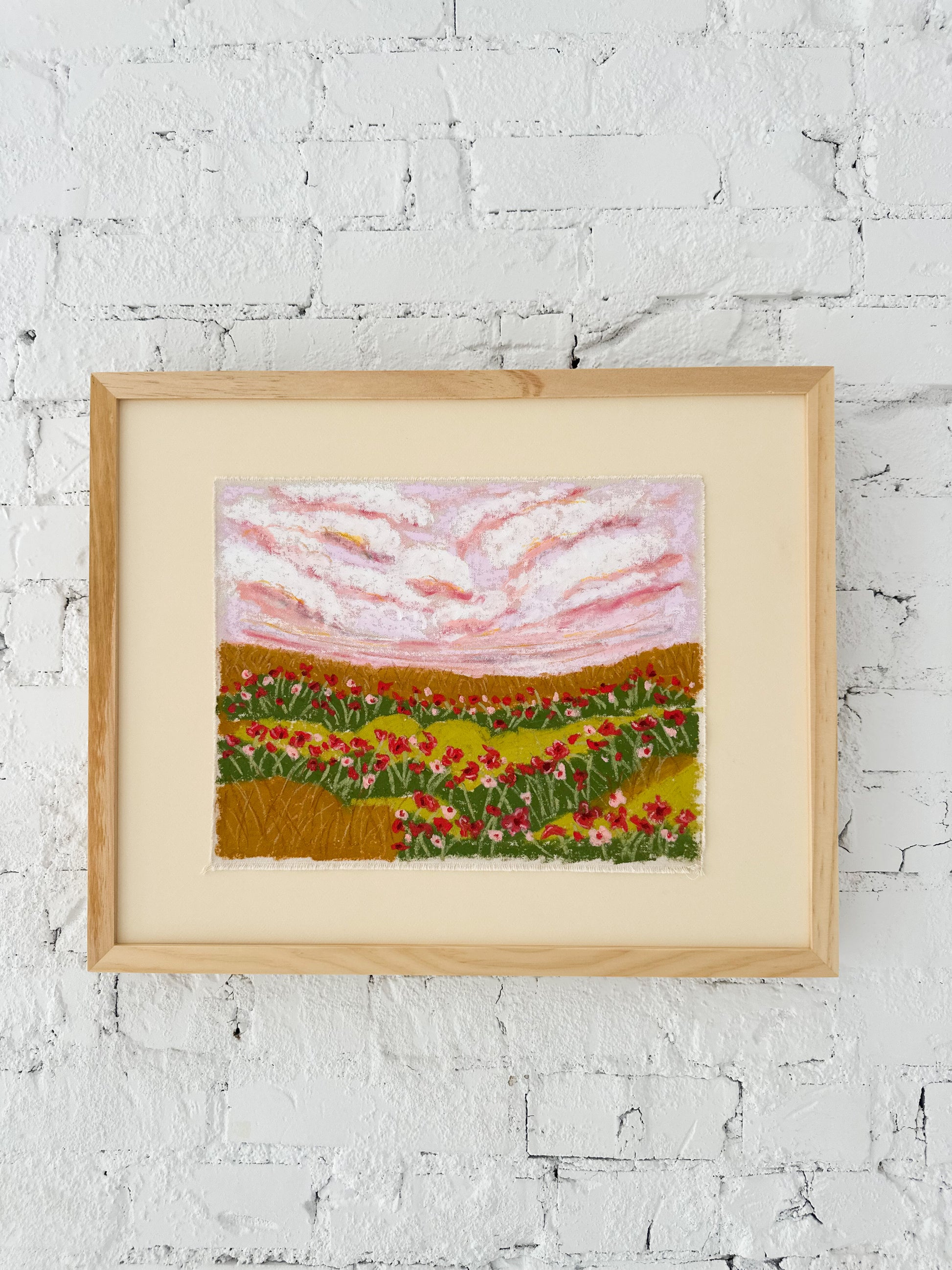 Framed artwork of a landscape with pink sky and green fields on a white brick wall.
