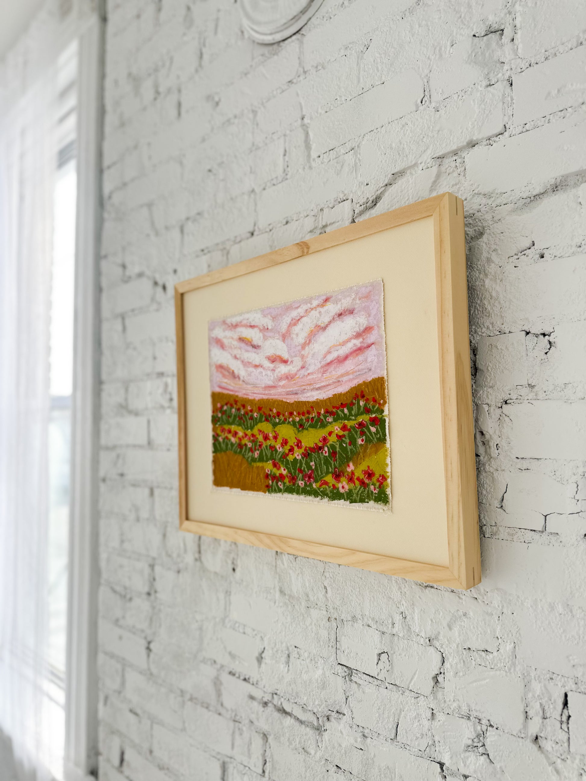 Framed artwork of a landscape with flowers on a white brick wall.
