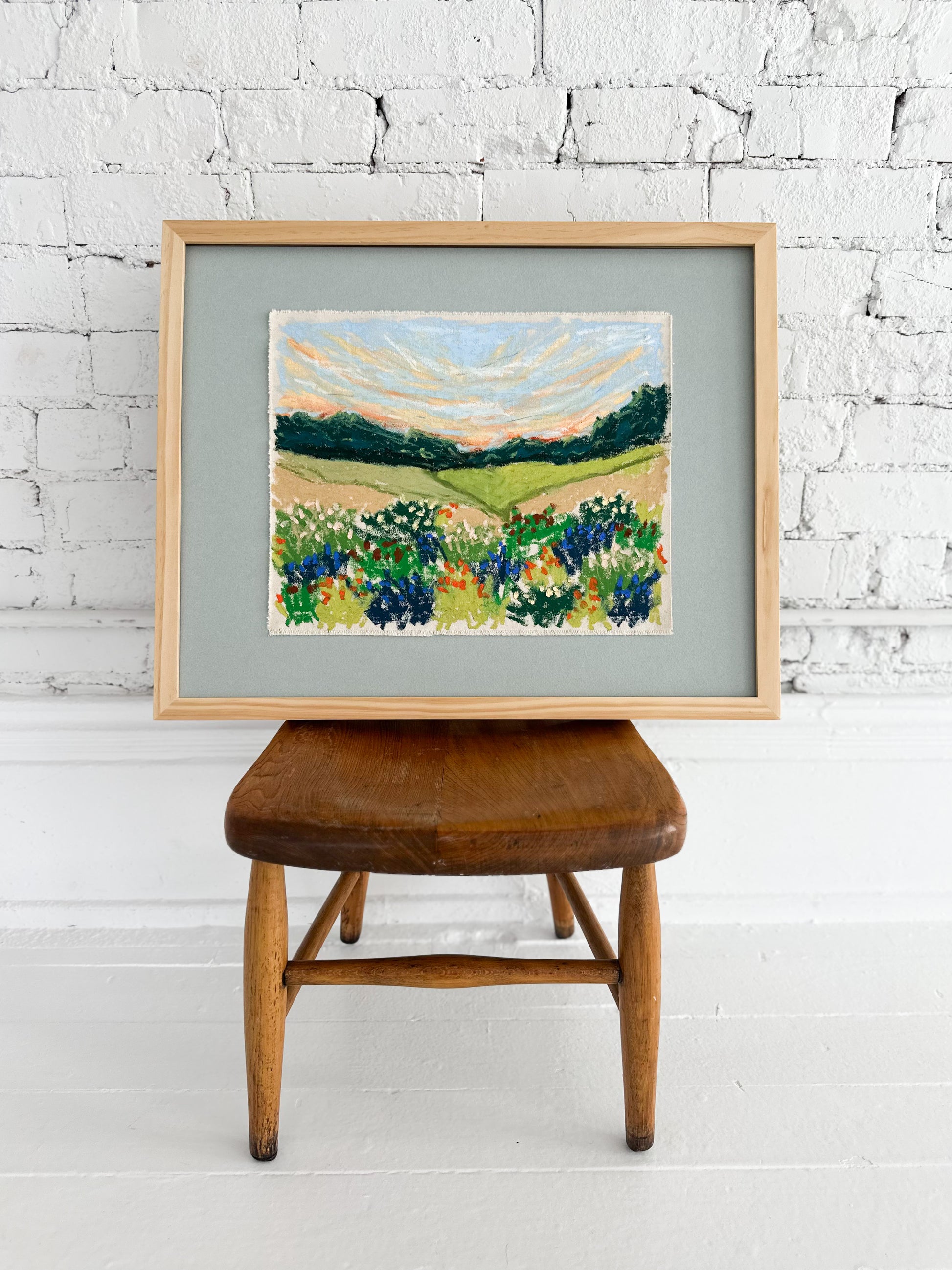 Framed painting of a landscape with flowers on a wooden stool against a white brick wall.