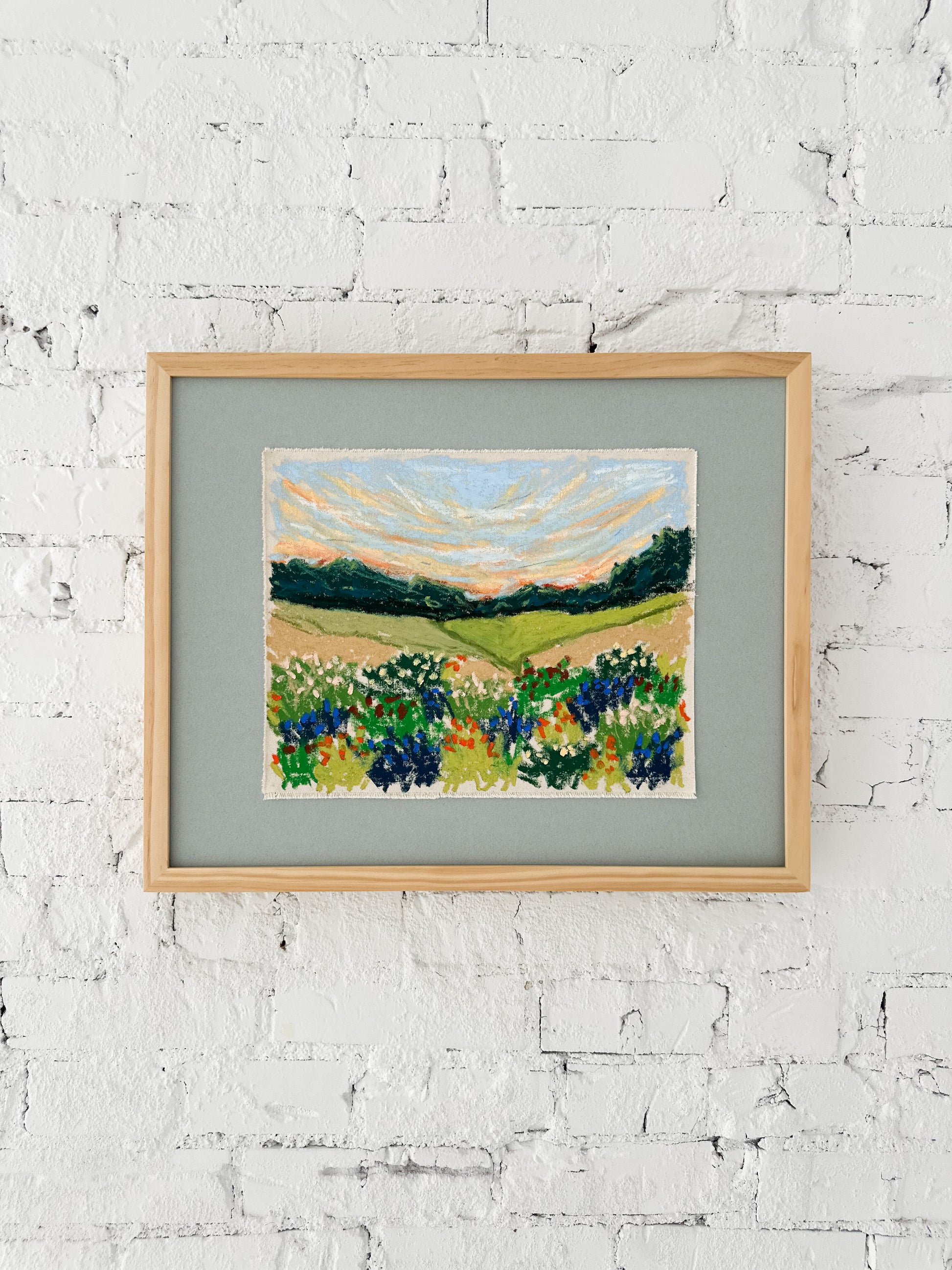 Framed artwork of a landscape with fields and trees on a white brick wall.