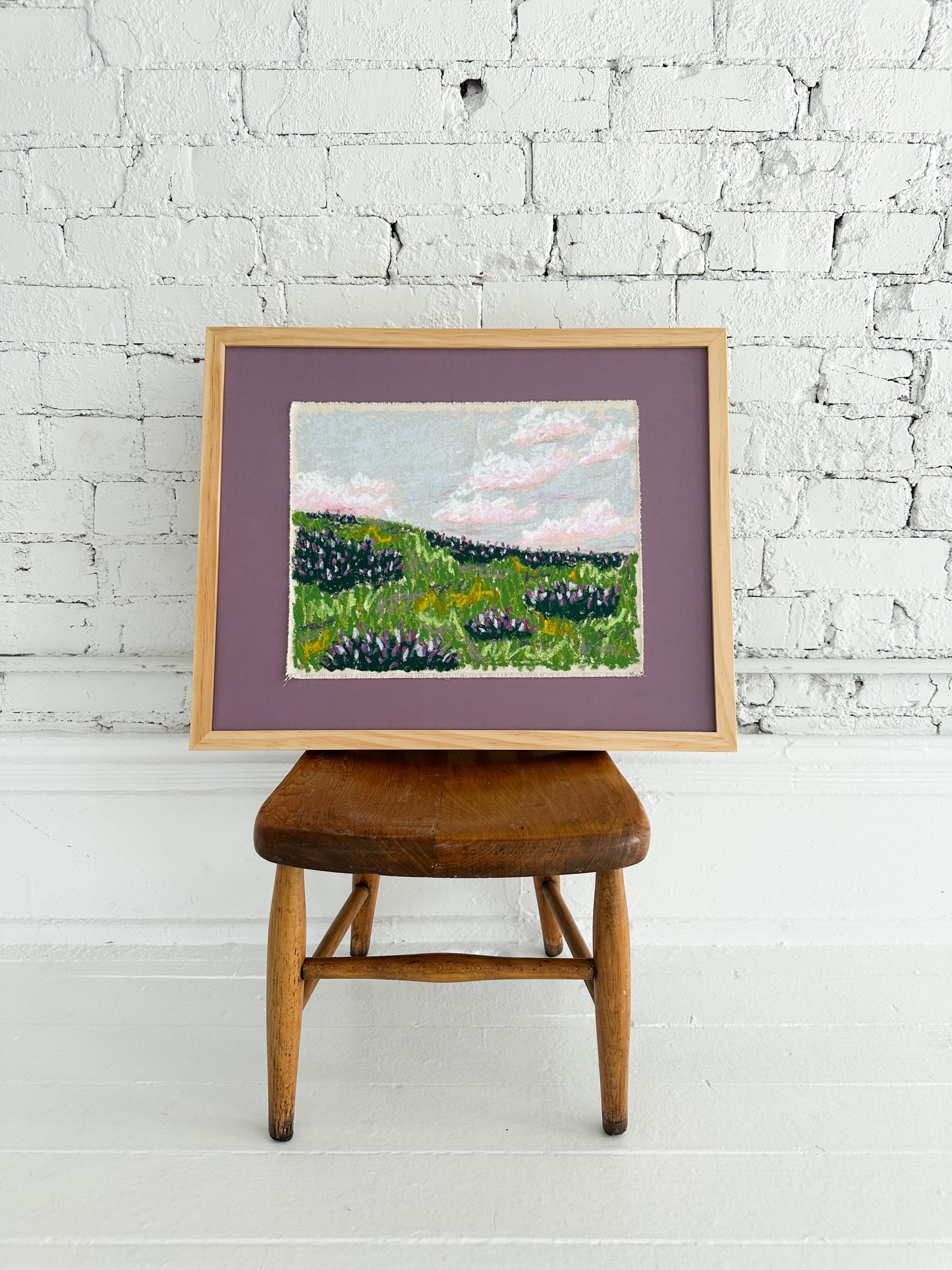 Framed painting of a landscape on a wooden stool against a white brick wall.