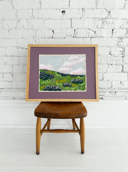Framed painting of a landscape on a wooden stool against a white brick wall.