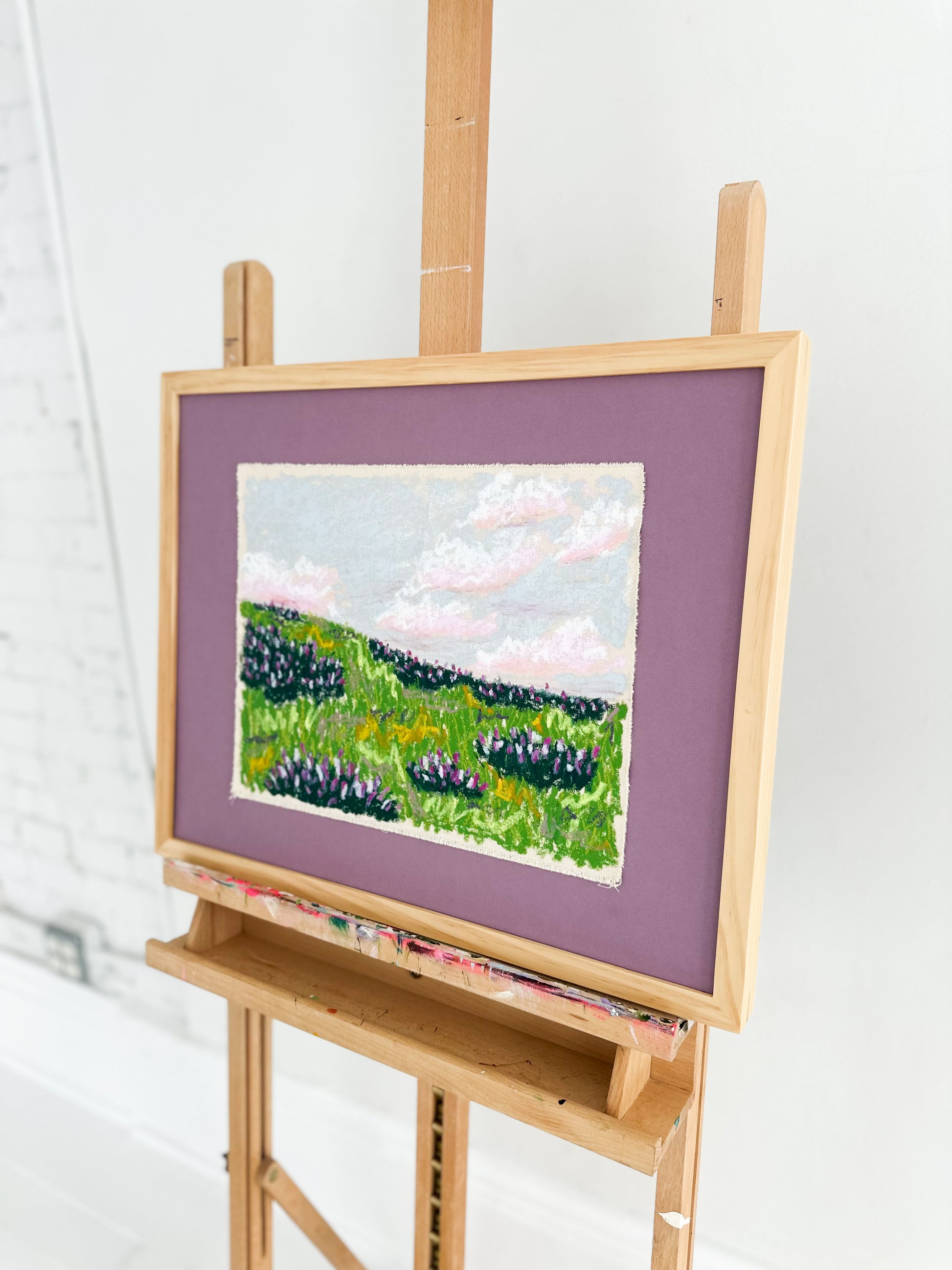 Painting of a landscape on an easel with a purple mat against a white background