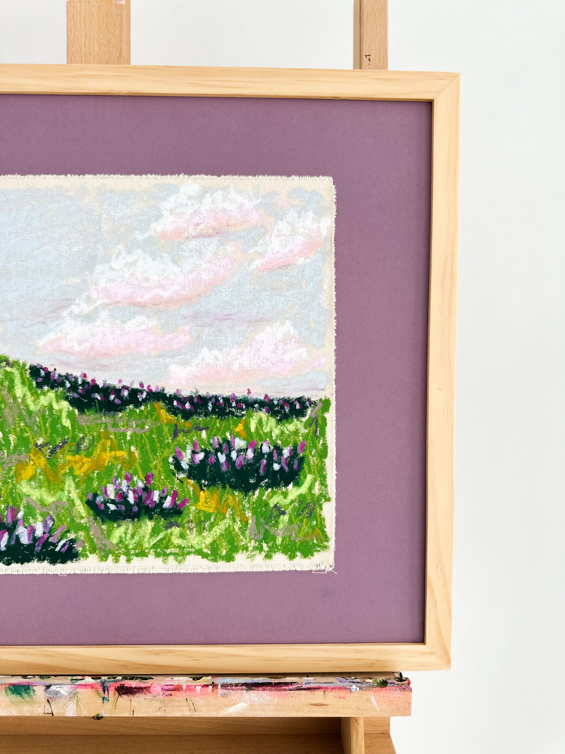 Framed artwork of a landscape with green fields and pink flowers on a purple background.