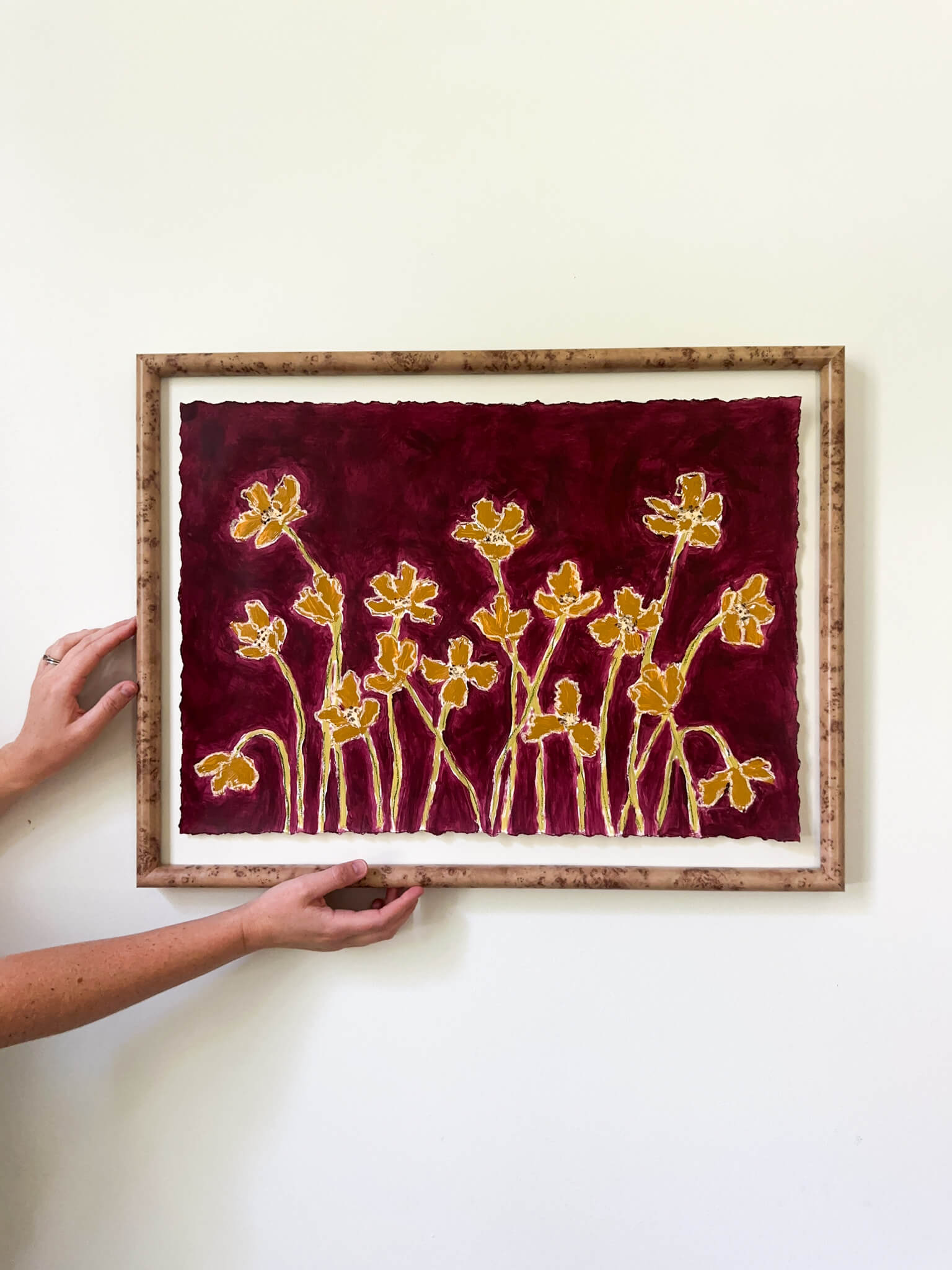 Framed artwork with yellow floral design on a red background, held by a person against a white wall.