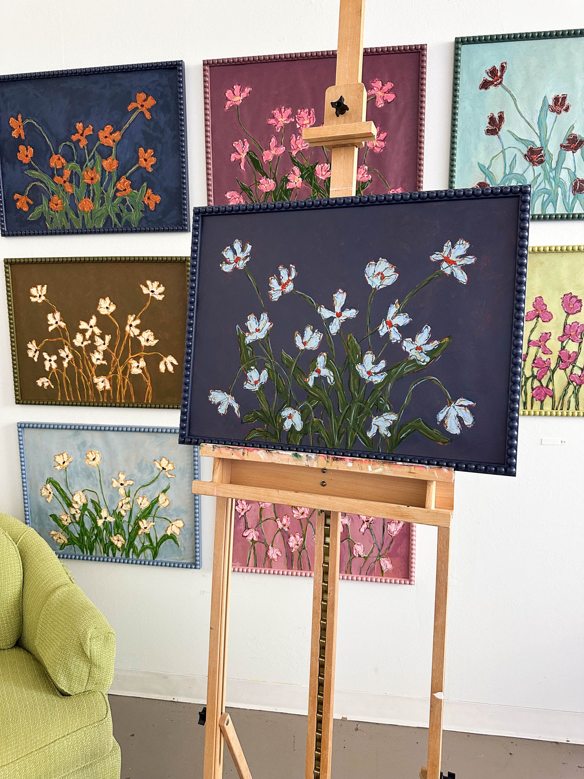 A painting of light blue flowers with green leaves on a dark blue background framed in a dark blue bobbin frame sitting on an easel in front of a white wall full of paintings.