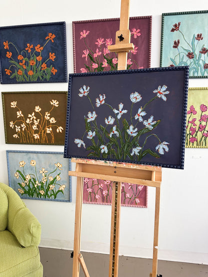 A painting of light blue flowers with green leaves on a dark blue background framed in a dark blue bobbin frame sitting on an easel in front of a white wall full of paintings.
