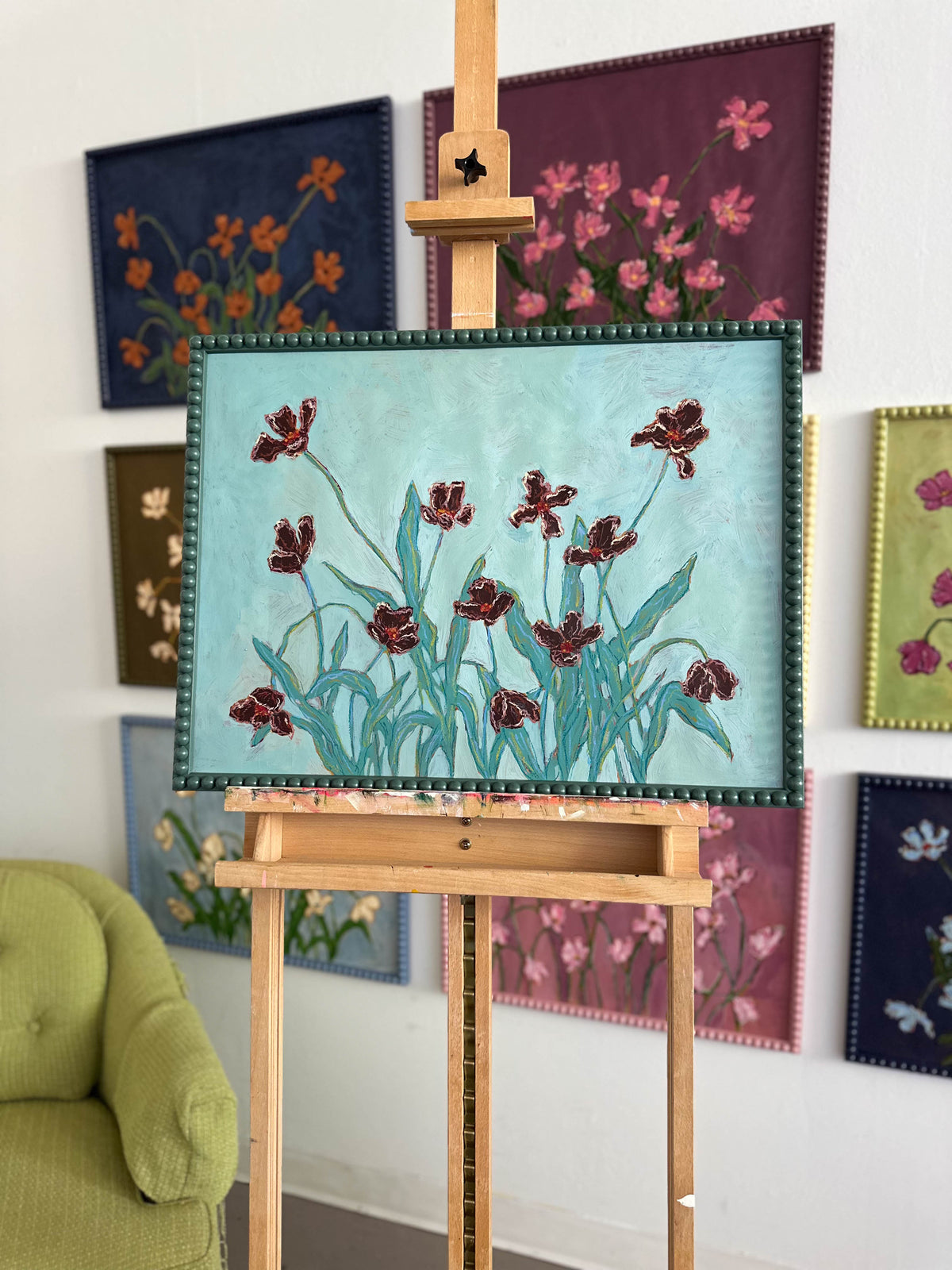 A painting of light pink flowers with green stems on a pink background and framed in a light pink bobbin frame sitting on an easel in front of a more painting hung on a white wall. 