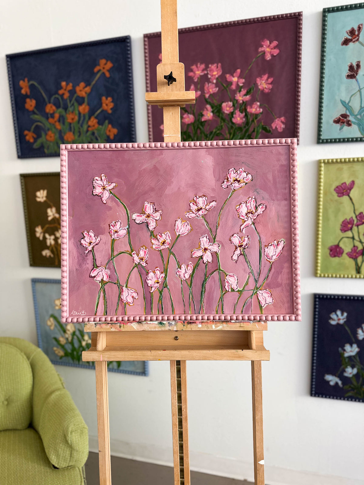A painting of light pink flowers with green stems on a pink background and framed in a light pink bobbin frame sitting on an easel in front of other paintings hung on a wall. 