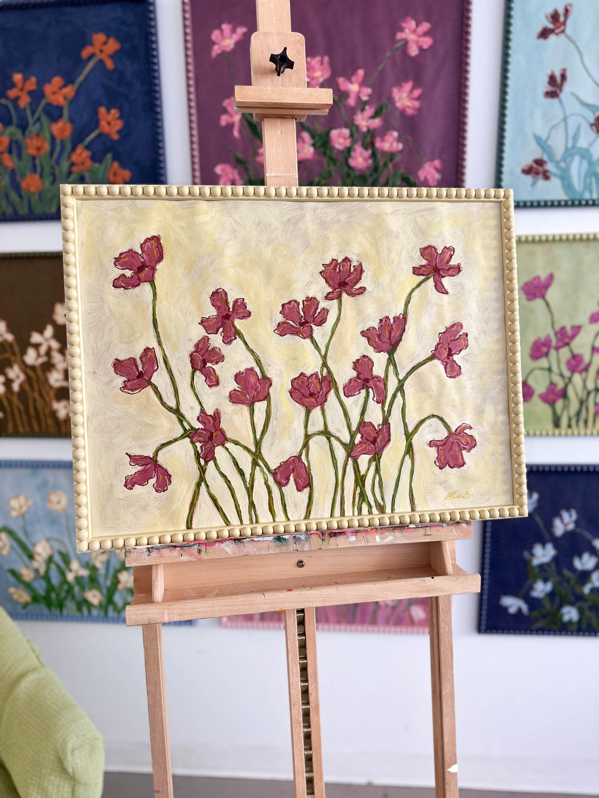 A painting of deep mauve flowers painting on a butter yellow background and framed in a butter yellow bobbin frame sitting on an easel in front of paintings hanging on a white wall.