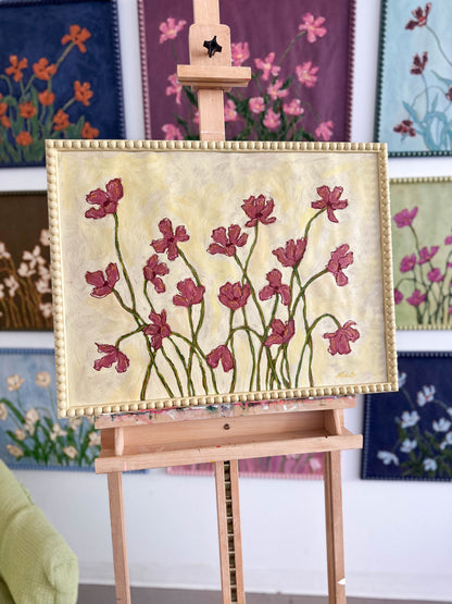 A painting of deep mauve flowers painting on a butter yellow background and framed in a butter yellow bobbin frame sitting on an easel in front of paintings hanging on a white wall.