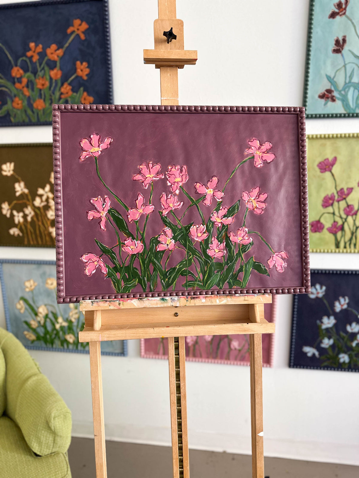 A painting of pink flowers with green leaves on a  plum background framed in a plum bobbin frame. It is sitting on an easel in front of a wall full on paintings.