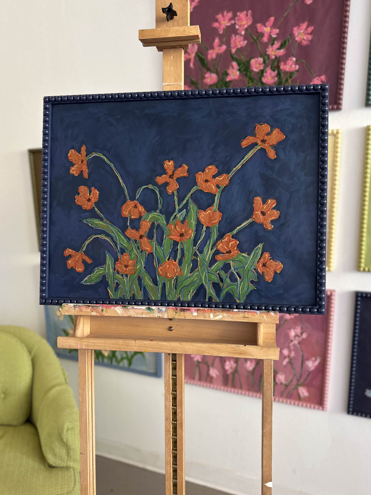 A painting of orange flowers with green leaves on a  deep blue background framed in a deep blue bobbin frame. The painting is sitting on an easel in front of a wall full of paintings. 