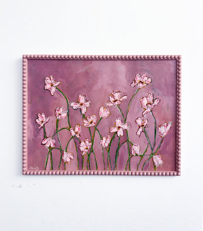 A painting of light pink flowers with green stems on a pink background and framed in a light pink bobbin frame. The painting is hung on a white wall.
