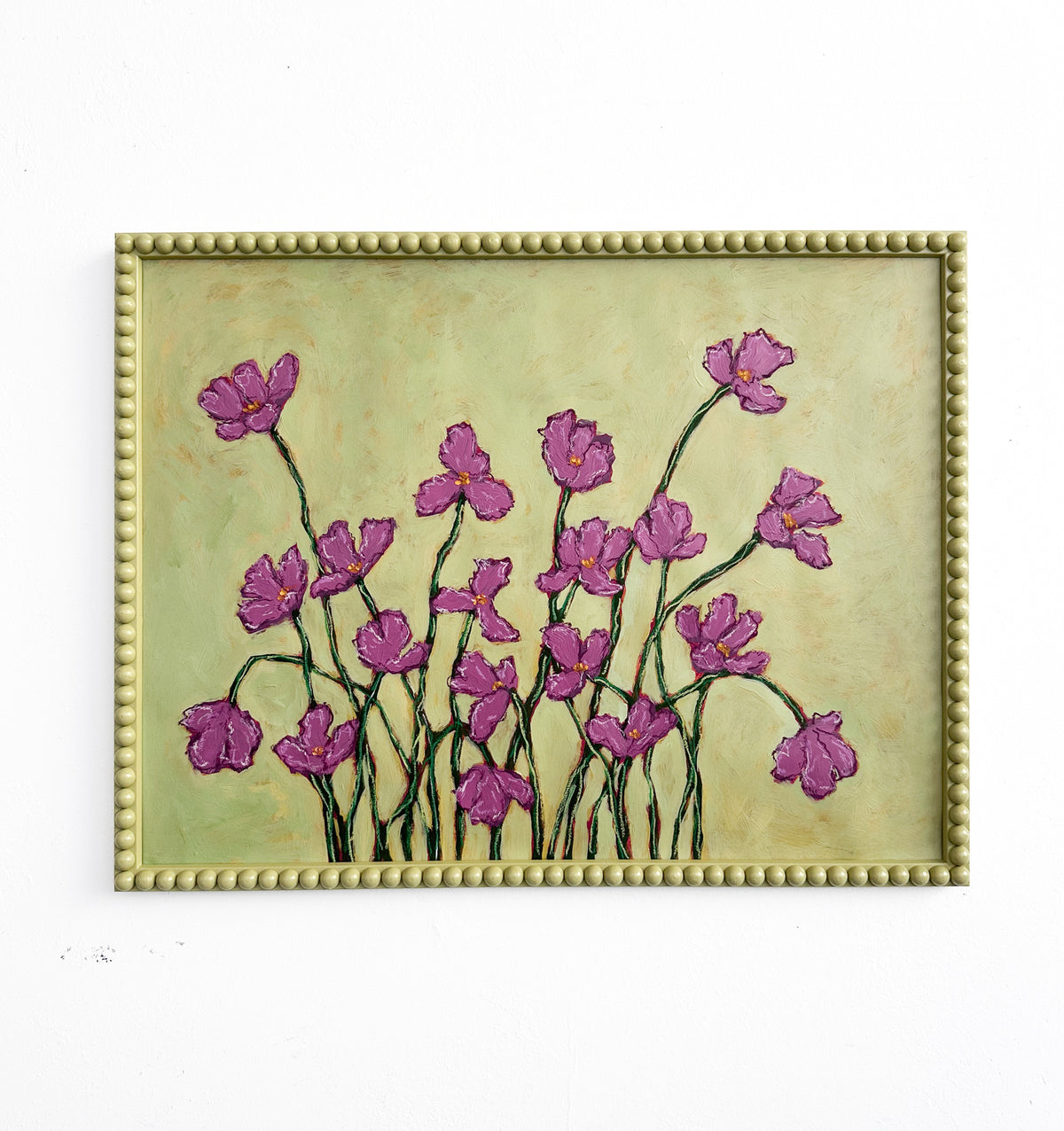 A painting of deep pink flowers with green stems  on a chartreuse background framed in a chartreuse green bobbin frame hanging on a white wall. 