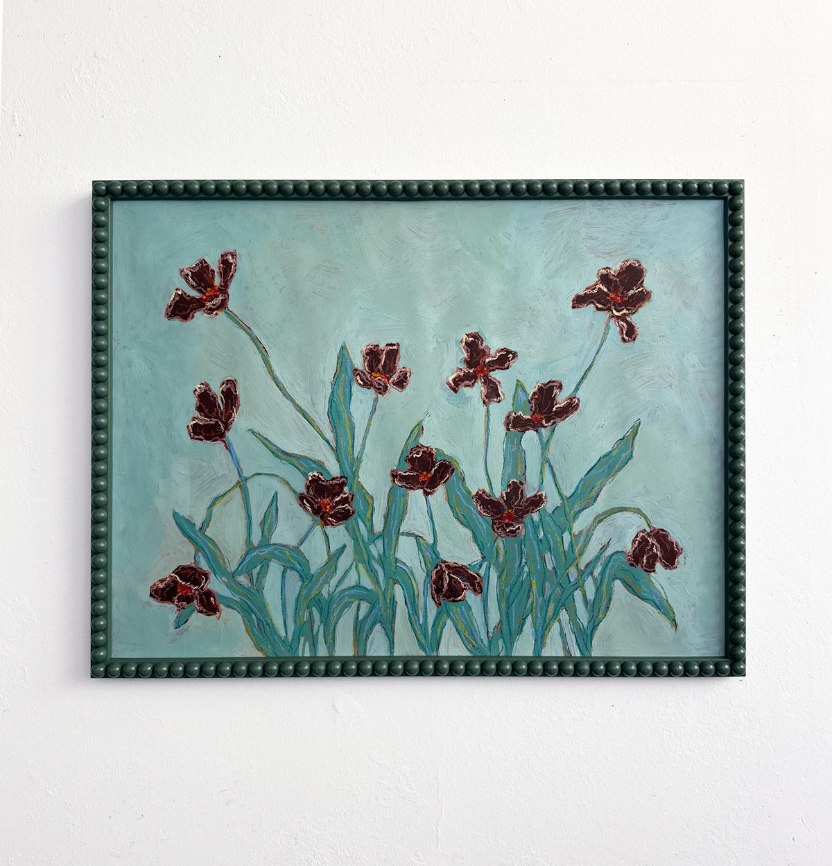 A painting of light pink flowers with green stems on a pink background and framed in a light pink bobbin frame. The painting is hung on a white wall.