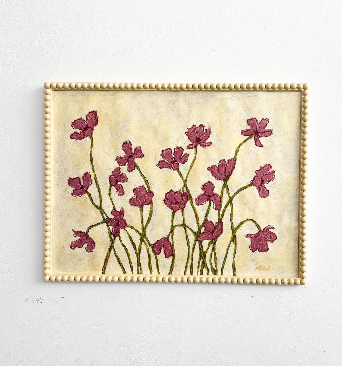 A painting of deep mauve flowers on a butter yellow background and framed in a butter yellow bobbin frame hanging on a white wall. 