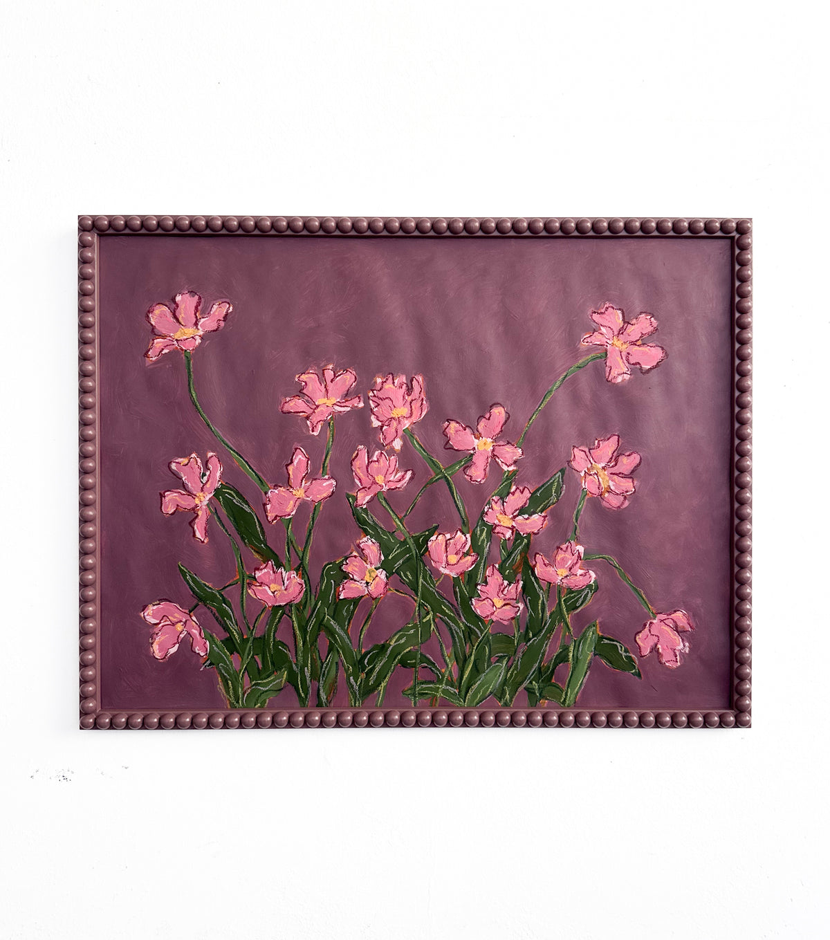 A painting of pink flowers with green leaves on a  plum background framed in a plum bobbin frame hanging on a white wall.