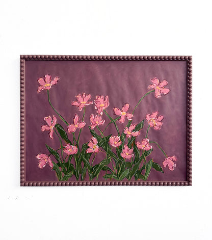 A painting of pink flowers with green leaves on a  plum background framed in a plum bobbin frame hanging on a white wall.
