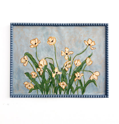 A painting of off white flowers with green leaves on a light blue background and framed in a blue bobbin frame. The painting is on a white background