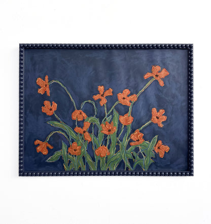 A painting of orange flowers with green leaves on a  deep blue background framed in a deep blue bobbin frame hanging on a white wall.