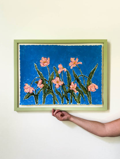 Framed artwork of pink flowers on a blue background, held by a hand against a white wall.
