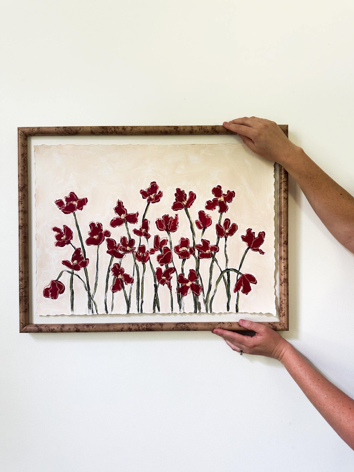 Framed artwork of red flowers on a white wall with hands adjusting the frame.